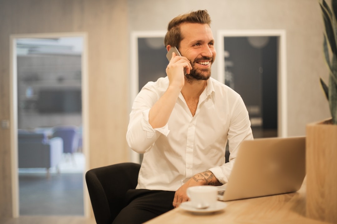 A man in white shirt taking a call on his mobile phone while working at a laptop