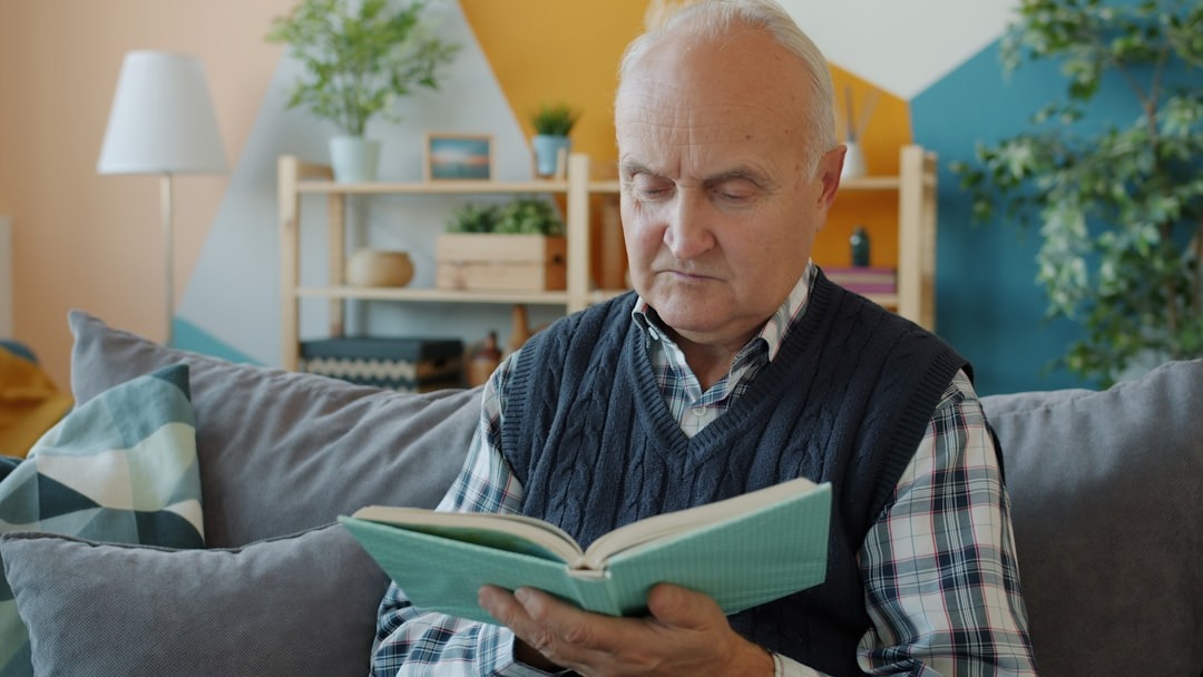Older man wearing check shirt and knitted v-neck gilet is sat on a sofa reading a book. Retirement Adviser UK